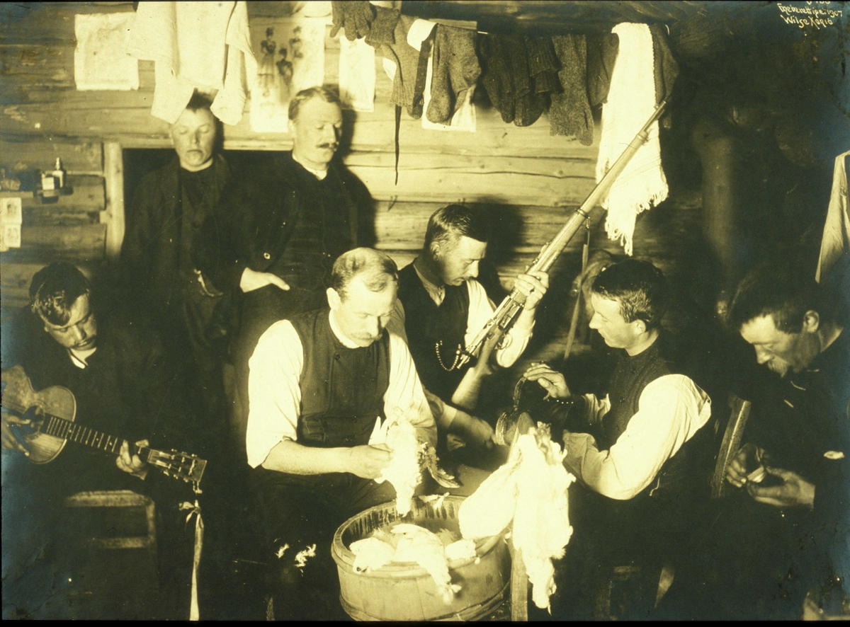 Påsketur til Ellevestølene, Filefjeld ved Helevann 1907. NF.W 06468.
Interiør  med klær til tørk under taket. 7 menn utøver forskjellige sysler, blandt annet gitarspill, sjekking av gevær og plukking av ryper.