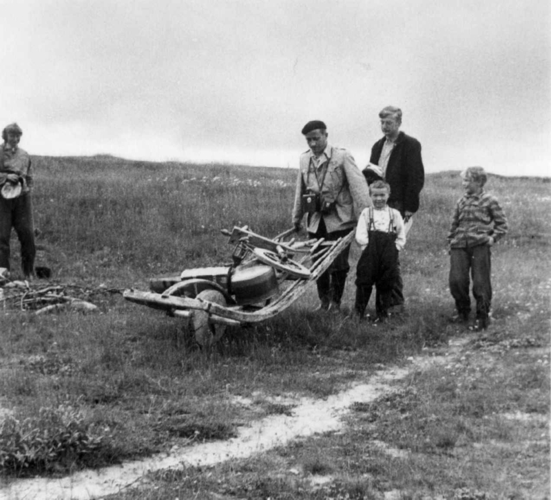 Dr. A. Nesheim med trillebår lastet med forskjellige gjenstander. Ved siden av går to barn og en voksen. Javrebainjarga 1952.